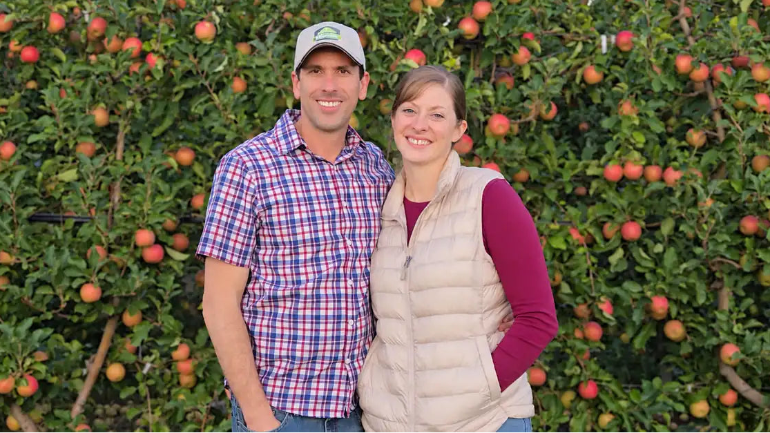 PollenPro founders Scott and Jen standing together in a Washington apple orchard surrounded by trees full of apples.